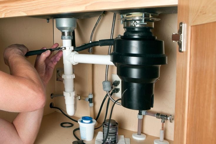 Plumber installing a garbage disposal beneath a kitchen sink in West Linn
